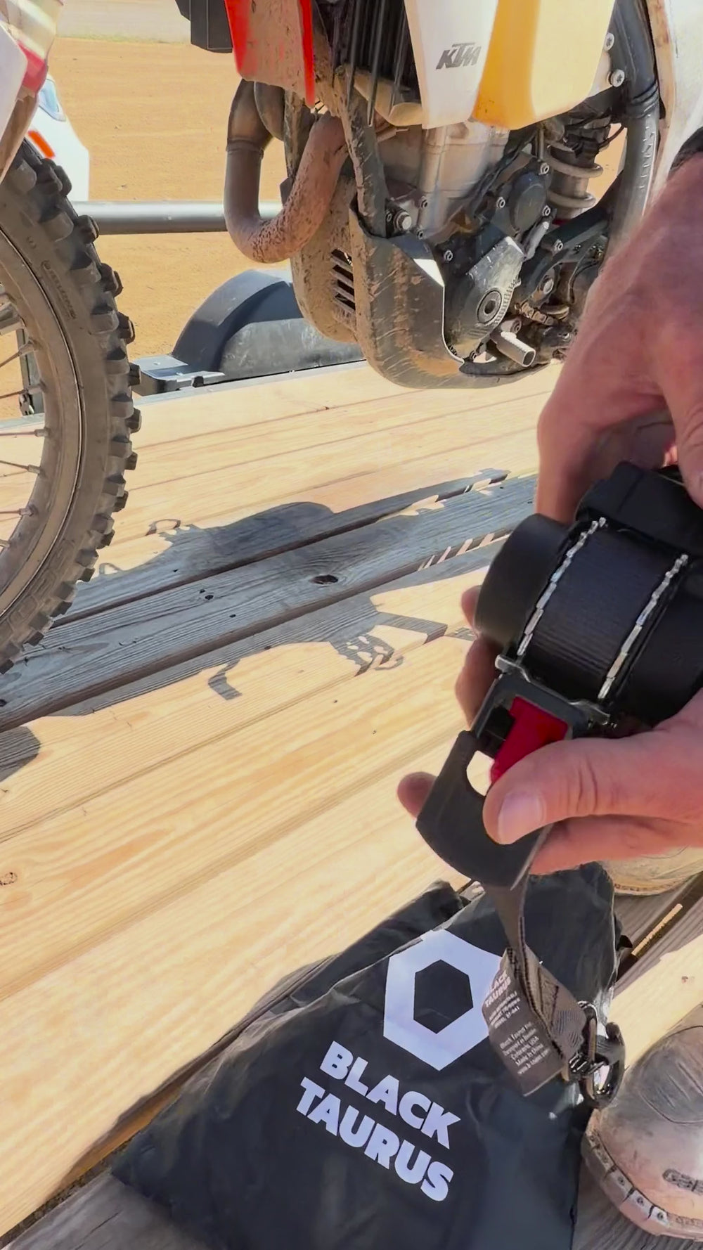Black Taurus Retract retractable ratchet straps securing a dirt bike on a trailer, with soft loops around the fork